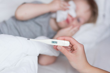 Close-up thermometer with fever heat 38. Mother measuring temperature of her ill kid at background. Sick child with high fever lying in bed at home. Mom Hand with thermometer close up