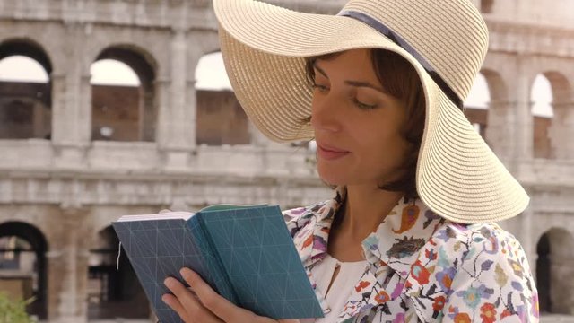 Young woman artist writes and draws on a notebook sitting at the table outside in front of the Colosseum in Rome. Elegant beautiful dress with large hat and colorful shopping bags.