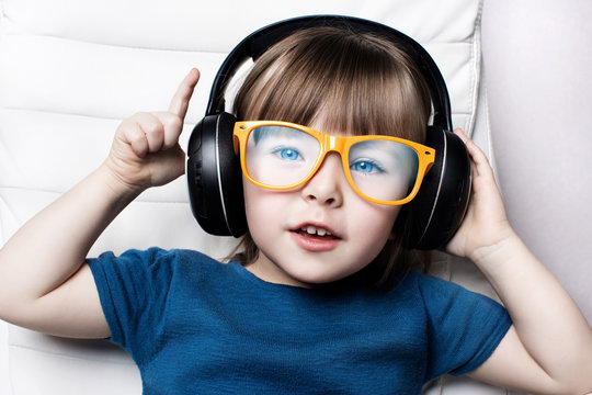A little girl in orange hipster glasses listens to music on headphones in the armchair of the house.