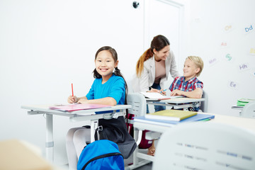 Happy schoolgirl looking at camera by desk with her teacher and classmate talking on background