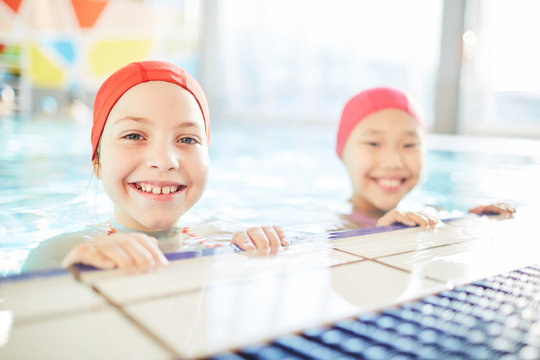 Cute smiling friendly girls in swim-caps looking at camera out of water