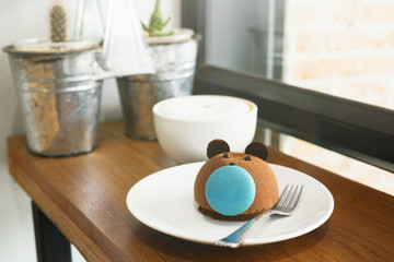 Fototapeta na wymiar Brown bear cake with blue macaron nose with cup of coffee on wooden table at cafe. Blurred plants and window in background. Text space