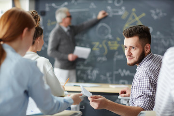 Student passing over folded paper with notice to one of his groupmates at lesson while teacher explaining formula by blackboard