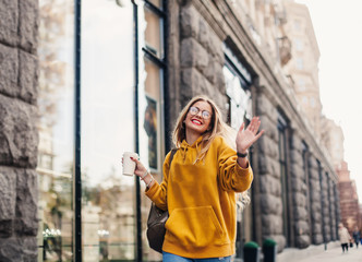 The concept of street fashion. young stylish girl student wearing boyfrend jeans, white sneakers bright yellow sweetshot.She holds coffee to go and dance . portrait of smiling girl in glasses