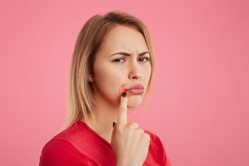 Discontent young beautiful female with unhappy look, has oral herpes, indicates at wound near lips, stands sideways against pink background. People, skin care and health problems concept. - obrazy, fototapety, plakaty