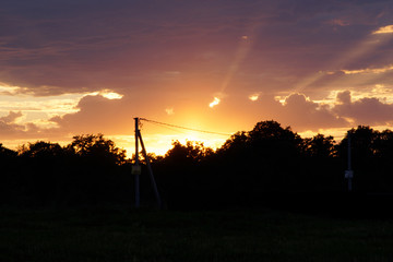 Sunset at the edge of the village. Against the background of a falling asleep light, a pole with electric wires, trees and wooden houses.