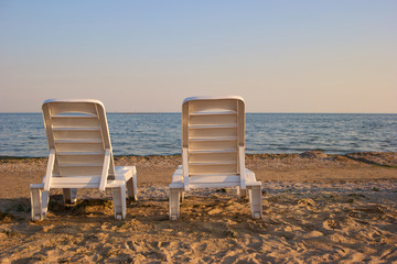 Back view empty chaise lounge before ocean. Rear view pair of beach chaise longues beside seashore. Two sunbeds on the beach: back view.