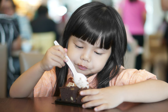 Asian Children Cute Or Kid Girl Enjoy And Fun With Happy Eating Delicious Brownie Chocolate Cake For Sweet Dessert Or Snack On Wood Table And Holding Spoon At Lunch In Restaurant Or Cafe