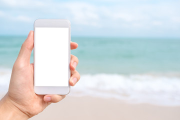 Mockup image of a hand holding and showing white mobile phone with blank black desktop screen in front of the sea and blue sky background