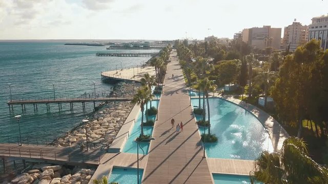 Aerial view of the coast of Limassol city in Cyprus. A view of the walk path surrounded by palm trees, pools of water, grass, the Mediterranean sea, piers, rocks and urban skyline.