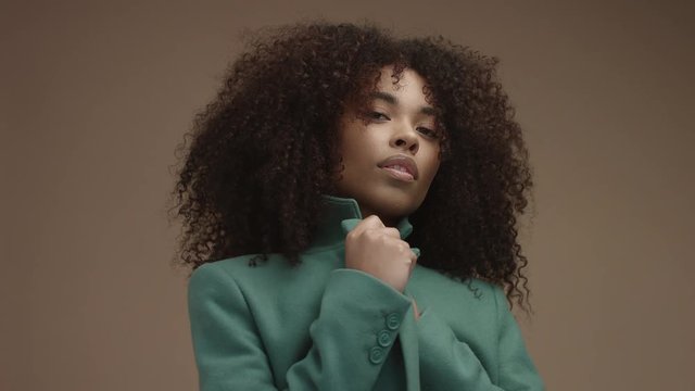 mixed race black woman in green coat in beige background with huge curly afro hair looking at camera and freezed from cold