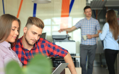 Two confident young people looking at touchpad while their colleagues working in the background.