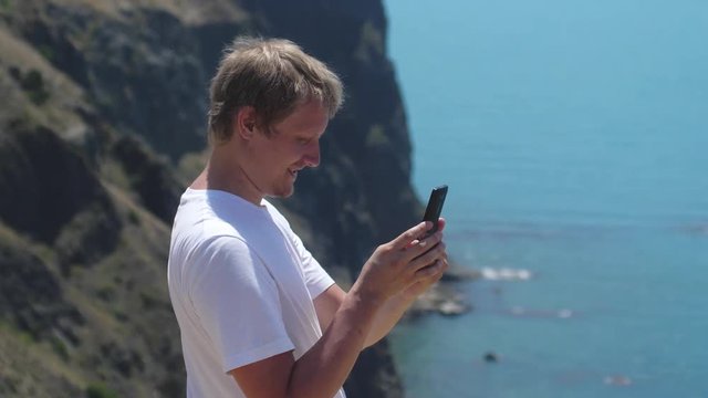 Young happy man taking photo with cellphone by the sea on the rock in slow motion. 3840x2160