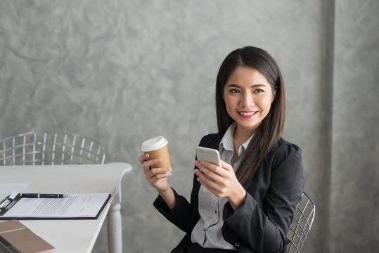 Asian business girl in her workstation at holding coffee cup and smartphone while smile