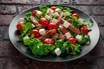 Juicy Beef Sirloin Steak Salad with roasted tomatoes, feta cheese and green vegetables in a black plate. healthy food