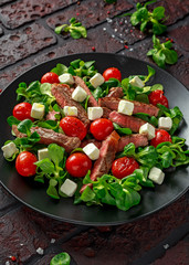 Juicy Beef Sirloin Steak Salad with roasted tomatoes, feta cheese and green vegetables in a black plate. healthy food