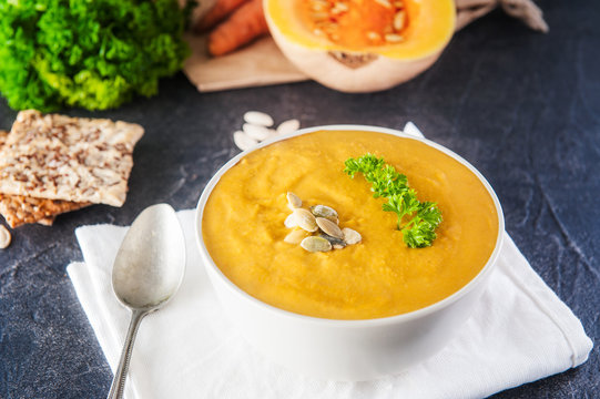 Bowl of Roasted pumpkin and carrot cream soup with parsley and pumpkin seeds on the dark stone served table with ingredients. Selective focus, Copy space.