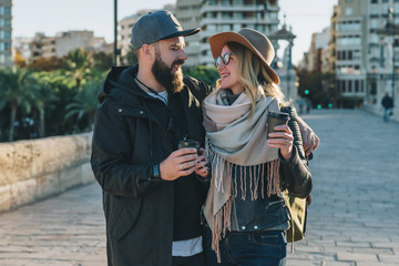 A couple of young tourists, friends are walking along the city street, drinking coffee while they are happily looking at each other. Vacation, travel, adventure, lifestyle. Blurred background.