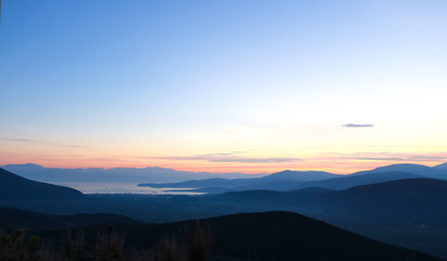 Sunset over blue layered mountains and the ocean far below with a few twinkling lights from houses and wind turbanes on a distant hill