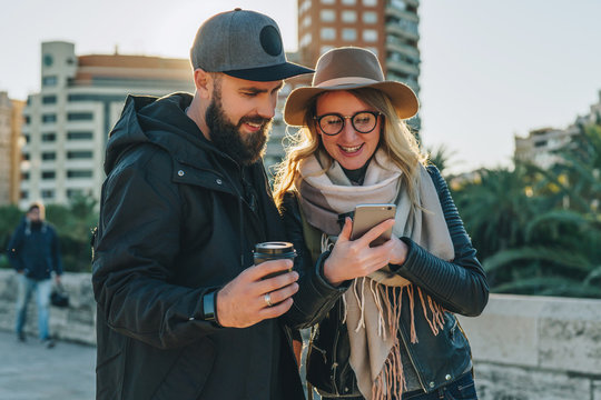 Couple of young travelers are standing on city street and using smartphone,holding cups of coffee.Girl shows guy an image on smartphone screen.Friends are walking around city.Lifestyle,social network.
