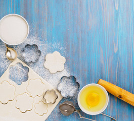 Raw puff pastry dough lies on a blue wooden table, sprinkled with flour. Figures for baking cut metal mold, sugar and egg in bowls