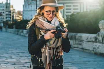 Fototapeta na wymiar Backlight. Young smiling woman tourist, photographer, hipster girl dressed in hat and eyeglasses, stands on city street and uses camera, looks images on screen. Vacation, travel. Blurred background.