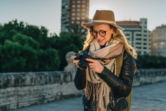 Backlight. Young smiling woman tourist, photographer, hipster girl dressed in hat and eyeglasses, stands on city street and uses camera, looks images on screen. Vacation, travel. Blurred background.