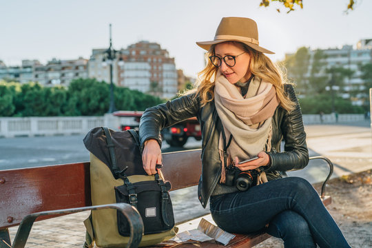 Young woman tourist, a hipster girl with camera, dressed in hat and eyeglasses, sits on bench in city street,is looking for something in her backpack. Vacation, travel, adventure.Blurred background.