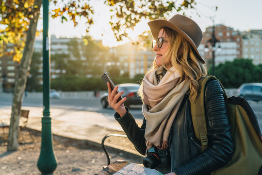 Young woman tourist, hipster girl with backpack, camera,dressed in hat, glasses,sits on bench in city street,using smartphone and holding map.In background,in soft focus,road and cars.Vacation,travel.