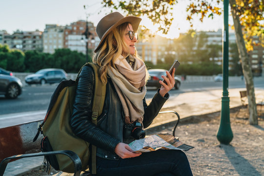 Young woman tourist, hipster girl with backpack, camera,dressed in hat, glasses,sits on bench in city street,using smartphone and holding map.In background,in soft focus,road and cars.Vacation,travel.