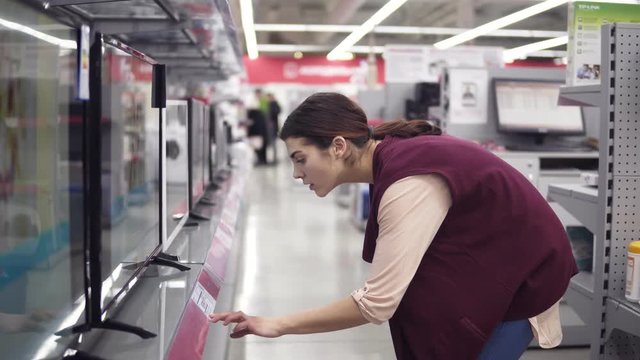 Young female costumer choosing new TV set compares pricetags for different TV screens in department of electronics in appliance store.