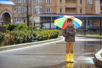 Little boy playing in rainy summer park. Child with colorful rainbow umbrella, waterproof coat and boots jumping in puddle and mud in the rain. Kid walking in autumn shower Outdoor fun by any weather