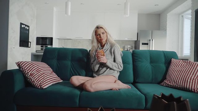 Gorgeous blonde woman sits on the couch holding a glass of water with fresh fruits. She is wearing wool sweater.