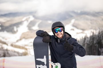 Fototapeta na wymiar Man snowboarder standing on the top of the snowy slope with snowboard, smiling to the camera, showing thumbs up at winter ski resort. Skiing and snowboarding concept