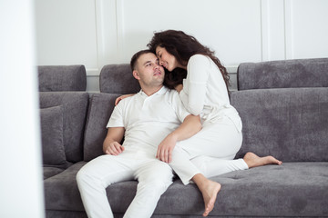 Obraz premium Loving young couple in white clothes hugging and relaxing on sofa at home. Love, family, happiness concept