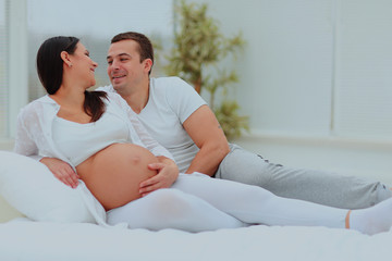 man looks lovingly at his pregnant wife lying on the white bed.