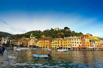Colorful buildings on the promenade of Portofino with boats floating in the marina and blue sky background