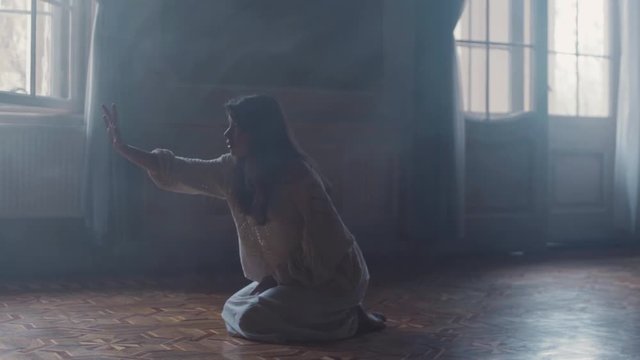 Young woman in long nighty sits on her knees in a dark room, she reaches out her hand and feels something in the air, scared she walks away. Paranormal activity, mystery