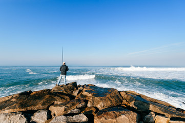 Fototapeta na wymiar Lonely fisherman looking farmale and fishing with spinning at atlantic ocean cape in sunny windy morning outdoors. Beautiful seascape. Fishing rod and gear. Saulty aquatic breeze. Hobby and leisure.