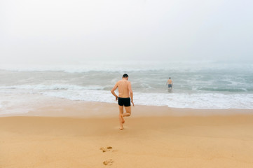 Topless man running from sandy beach in misty stormy ocean. Adult male enjoying swimming like child. Happy unrecognizable person. Water procedures. Healthcare and recreation. Crazy guy on vacation.
