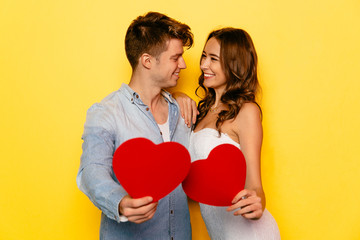 Fototapeta na wymiar Cheerful smiling young couple celebrating St. Valentine's day, holding red hearts, looking to each other. Yellow background.