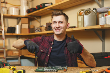 Multimeter for electrician. Caucasian young man in plaid shirt, black T-shirt digital electronic engineer repairing, soldering computer PC motherboard in workshop at wooden table with different tools.