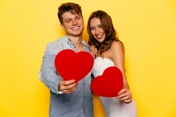 Perfect beautiful couple celebrating St. Valentine's day, holding red hearts, looking at camera. Yellow background.