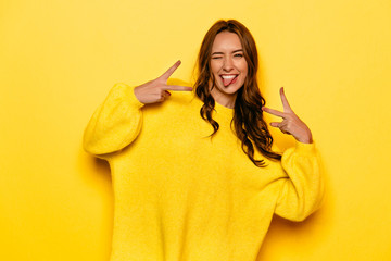 Smiling pretty girl with curly hair winking, showing a tongue, peace gesture. On yellow background.