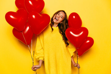 Young attractive girl with long curly hair, in yellow sweater holding red air balloons, posing at...