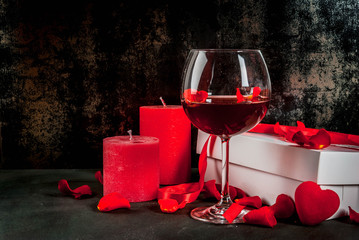Valentine's day concept, white wrapped gift box with red ribbon, rose flower petals, red wine glass, with red candle, on dark stone background, copy space