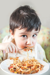 Child eating spaghetti with vegetables. Kid having fun eating. Brown haired boy with face covered in sauce. Weekend, warm and cozy scene in the kitchen.