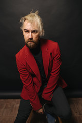Obraz premium Red jacket and wind in blond hair. Portrait of masculinity. Portrait of handsome young bearded man looking at camera while standing against black background