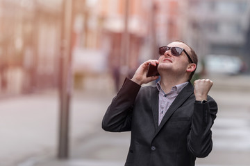 Young entrepreneur businessman talking on his smart phone, scores a big deal and smiles, outdoors in the streets