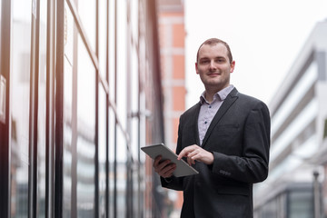Fototapeta premium Young entrepreneur businessman looking and smiling, checking and reading his email messages on his tablet, outdoors in the streets